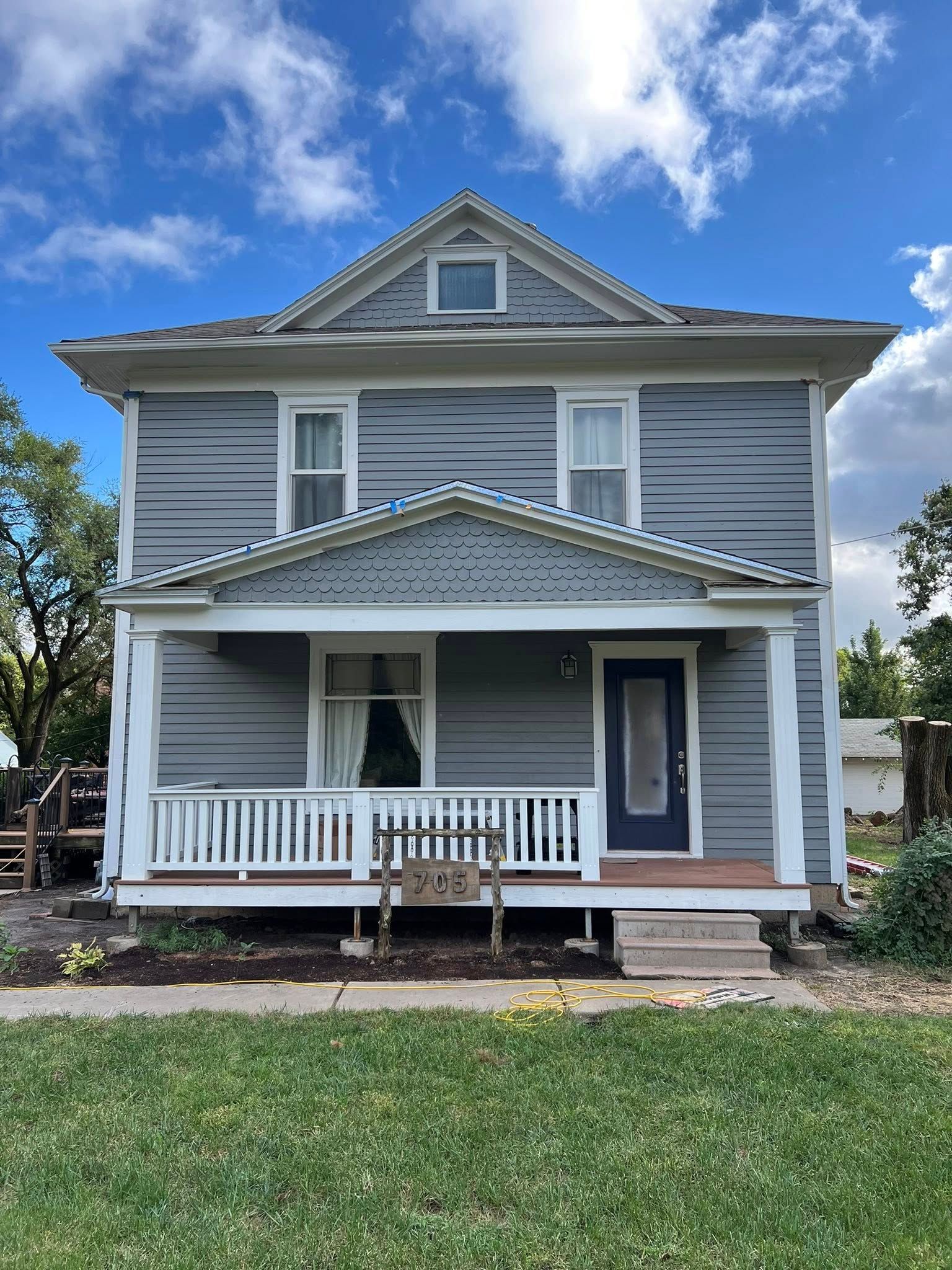 Two-story house with gray siding, white trim, and a porch, set against a blue sky with clouds.