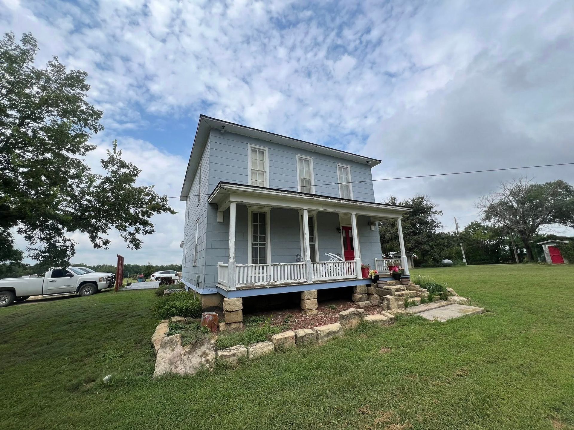 Two-story blue house with porch, in a grassy yard, under a cloudy sky. A truck is parked to the left.