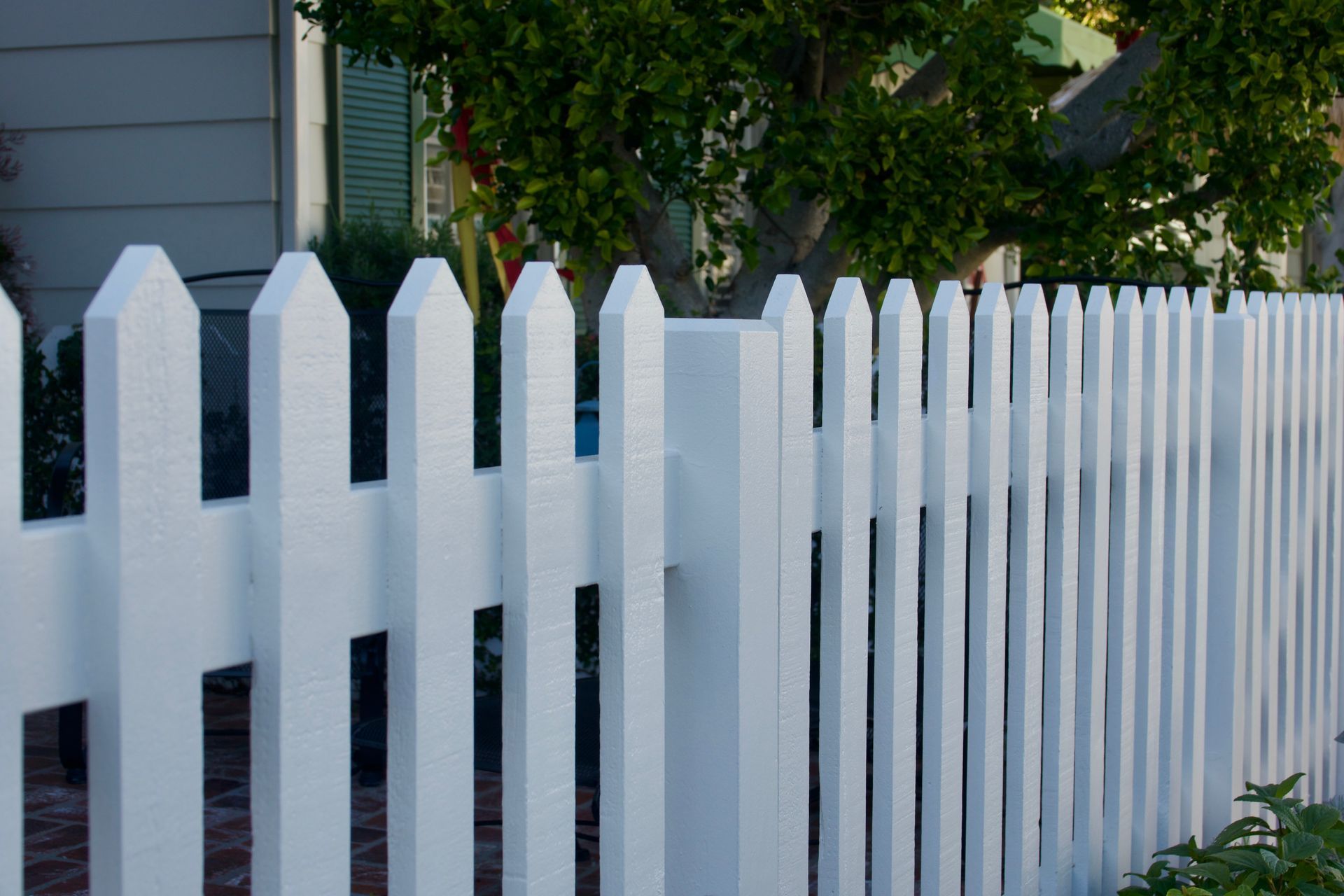 White picket fence in front of a house, with green tree in the background.