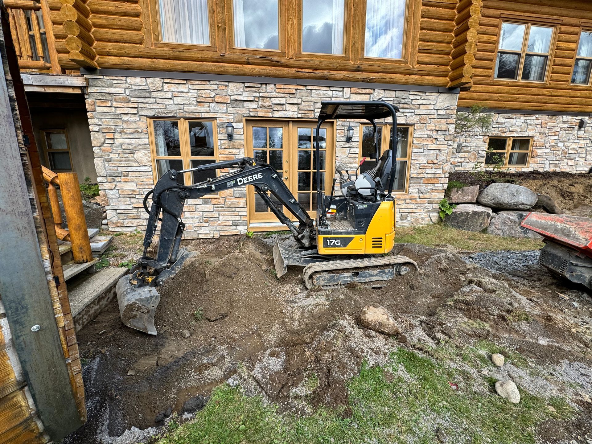 Une petite excavatrice creuse un trou devant une cabane en rondins.