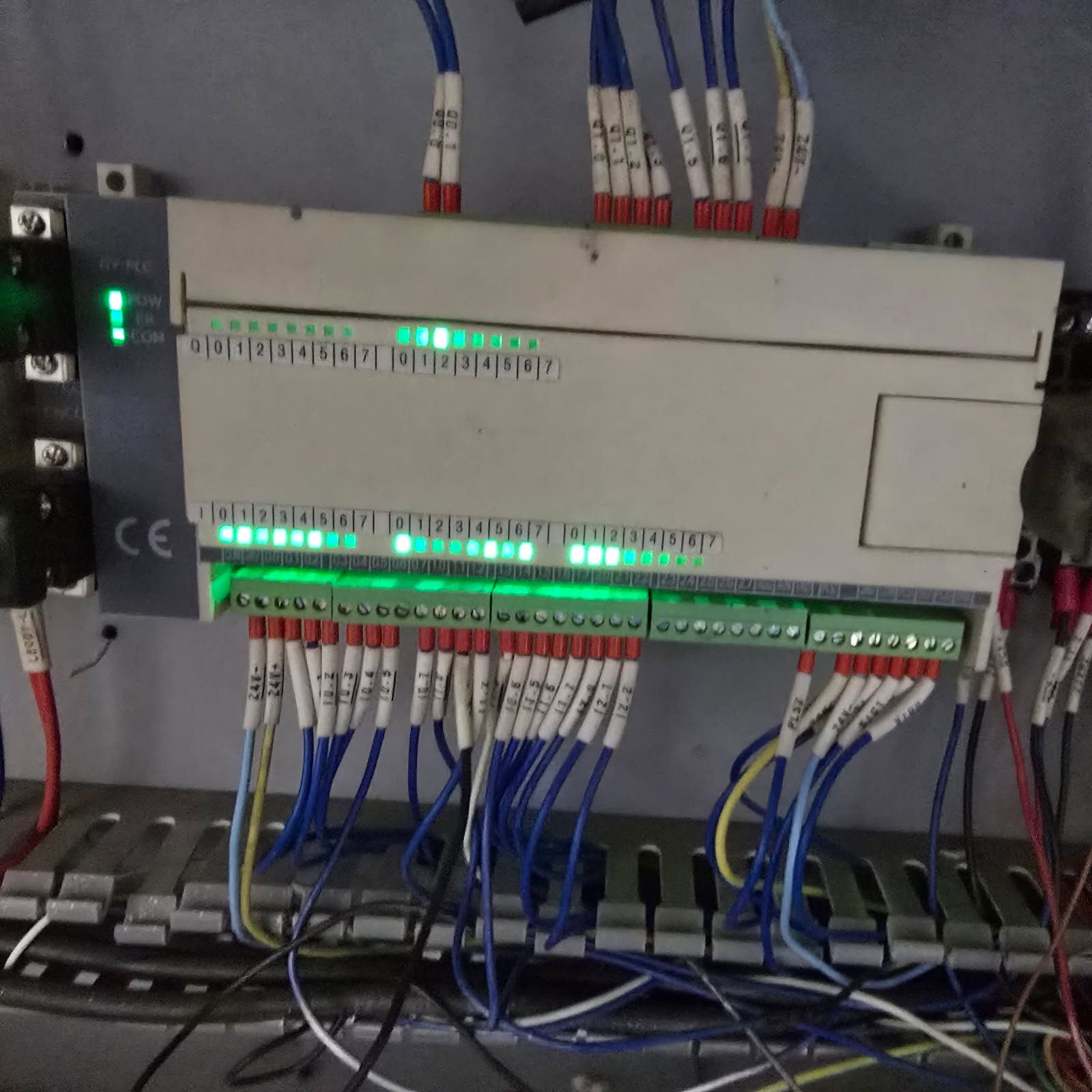 A rectangular PLC controller mounted on a panel, featuring multiple green status indicator lights and wired connections.