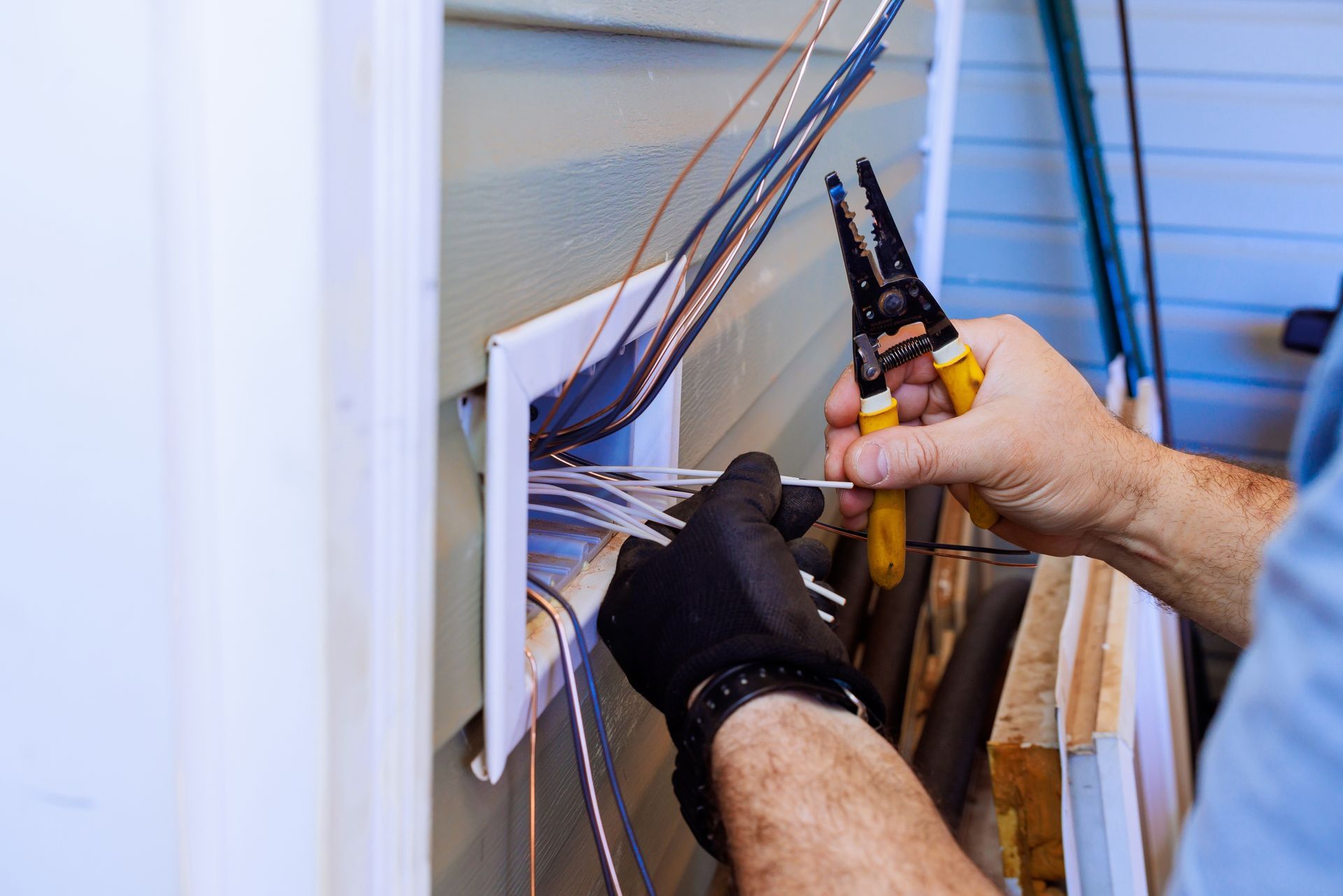 A person wearing gloves using wire strippers to work on electrical wiring protruding from a wall.