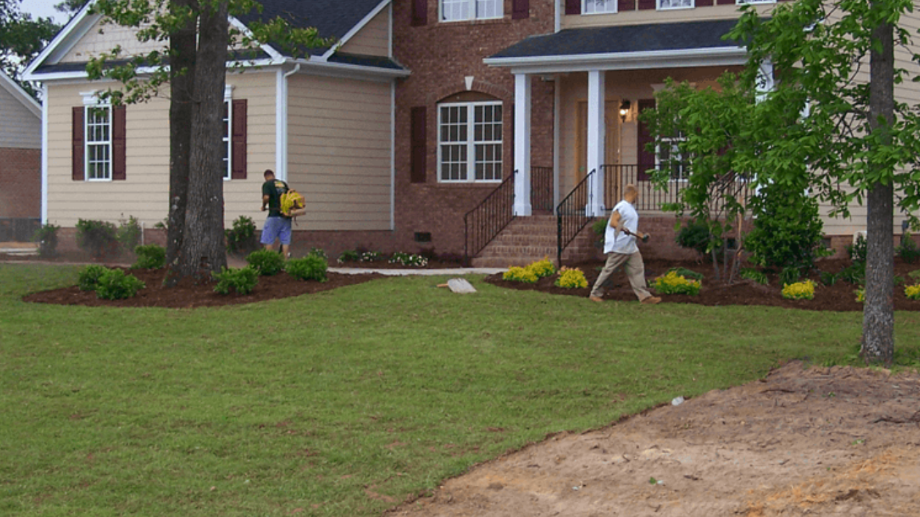 Two people spraying a lawn in front of a brick and tan house in Fayetteville, NC.