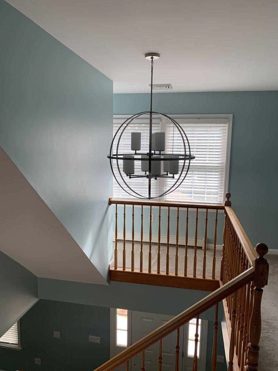 A two-story entryway with a blue wall and brown railing. A black chandelier hangs above.