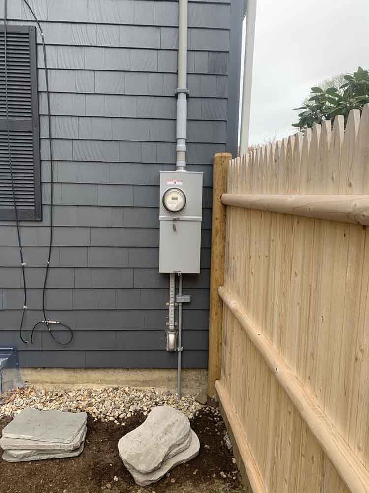 Gray electrical box with meter mounted on gray house siding next to a wooden fence.