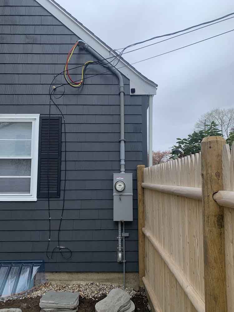 Gray house exterior with electrical meter, conduit, and wiring running to the roofline. Fence and window are visible.
