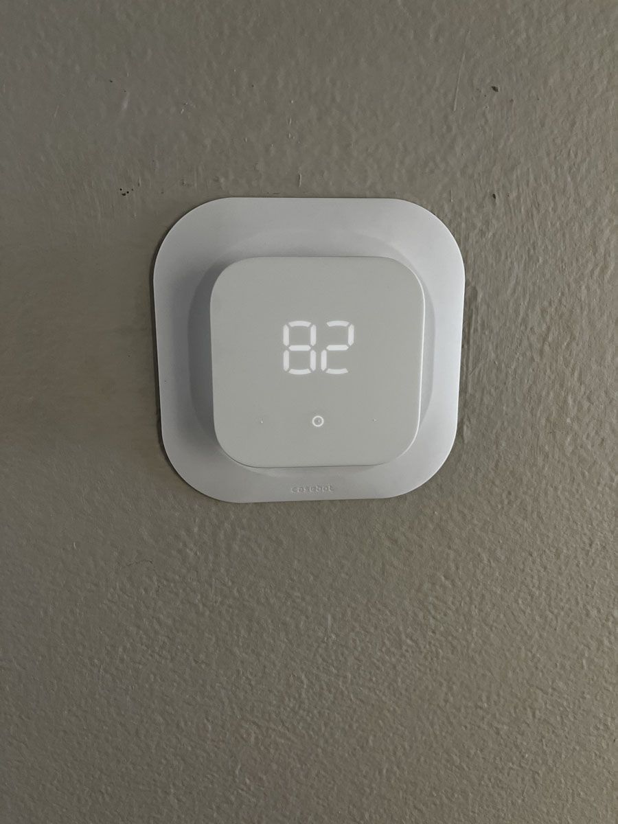 White smart thermostat displaying 82 degrees, mounted on a gray wall with a square white base.