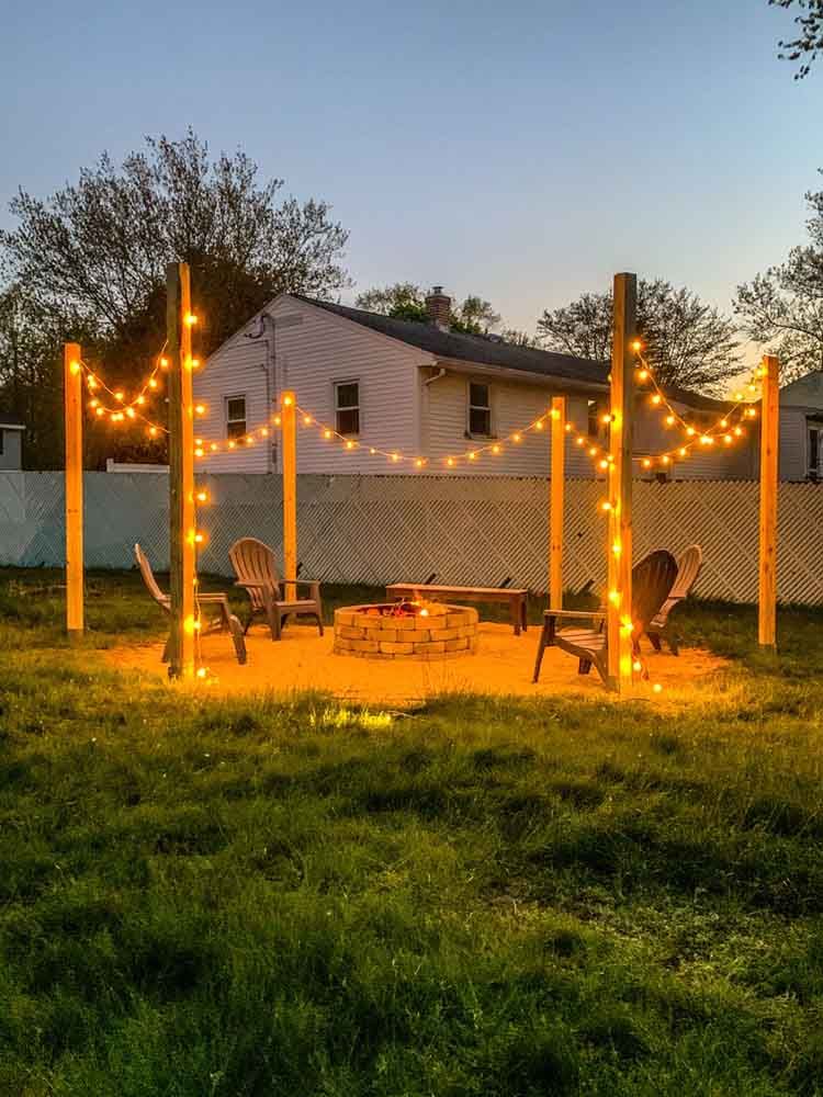 A backyard fire pit area lit with string lights and wooden posts. Chairs face the pit, near a house at dusk.