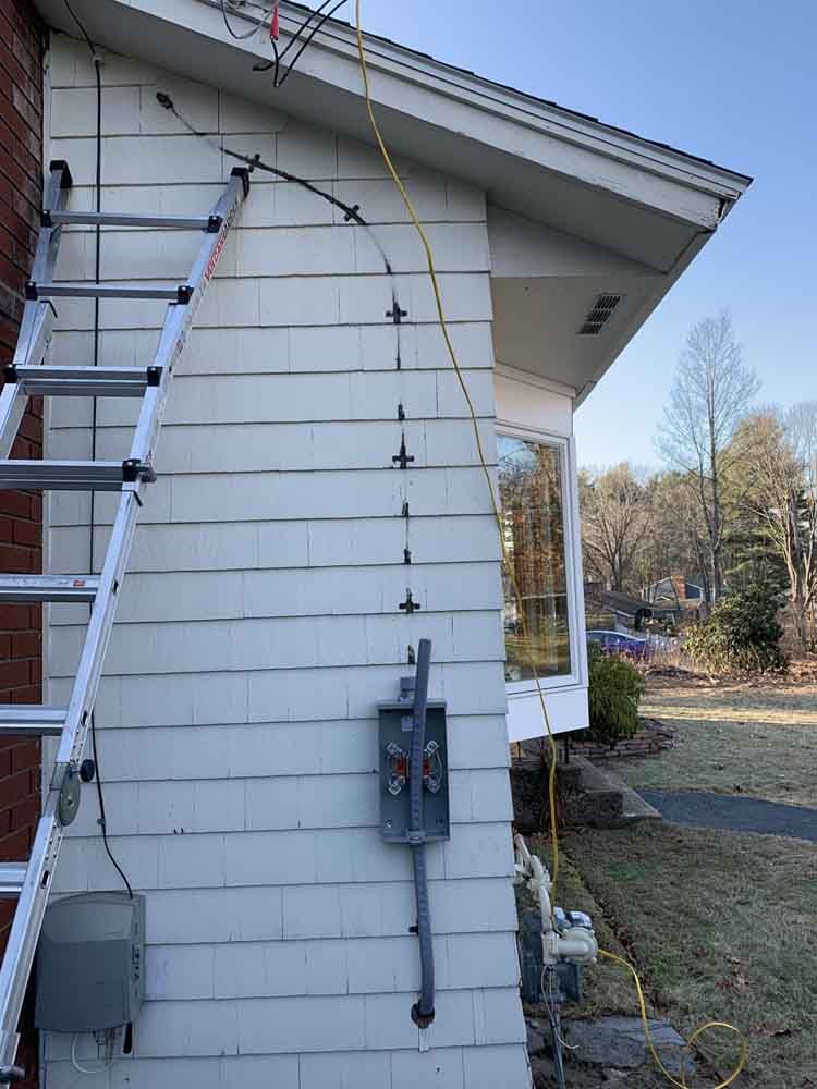 Ladder against a house with wiring along the siding. A yellow cord runs down to a grey box.