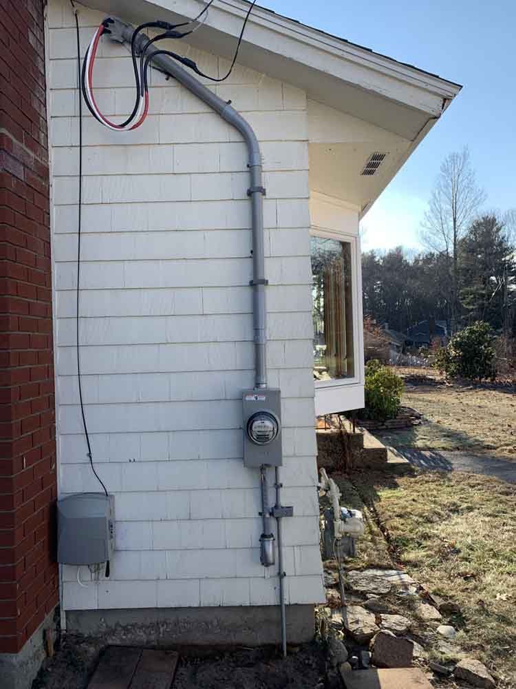 Electrical meter and conduit on the side of a white house with a brick chimney.