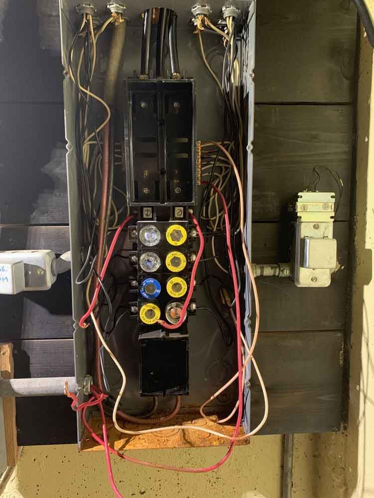 Open electrical fuse box with exposed wires and fuses against a dark wooden wall.