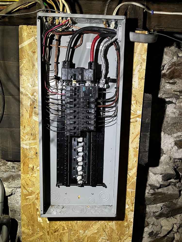 Electrical panel with numerous wires and circuit breakers, mounted on a wooden surface.