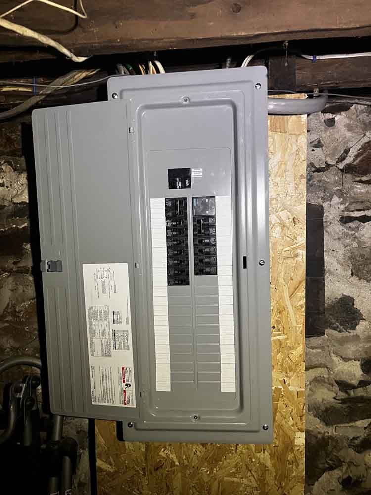 Electrical panel with the door open, mounted on wood against a stone wall.