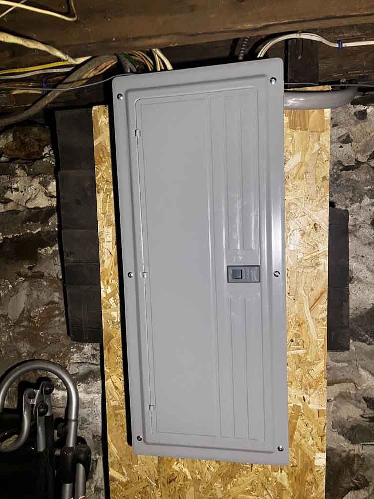 Electrical panel in a basement setting, mounted on plywood. Gray panel with closed door and breaker switch.
