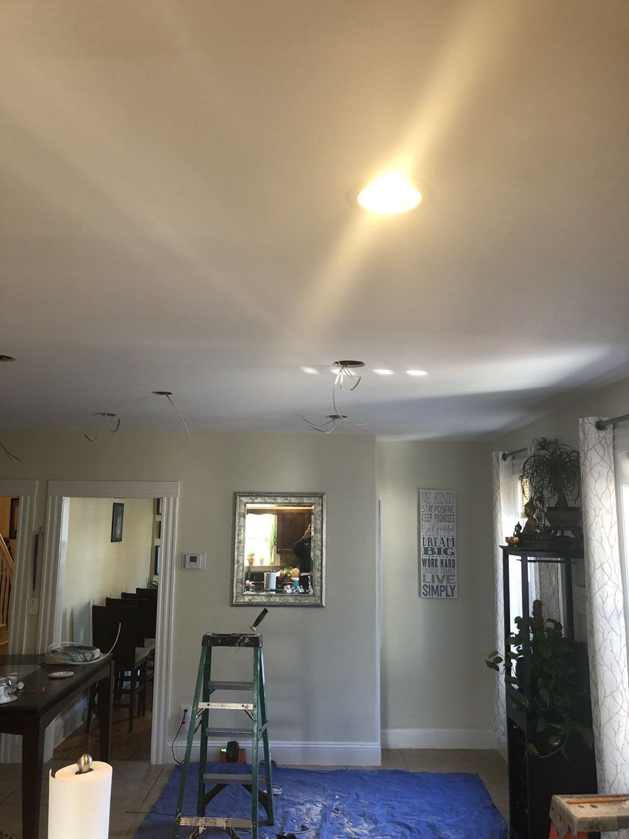 A person on a ladder is working on ceiling lights in a room with pale walls.