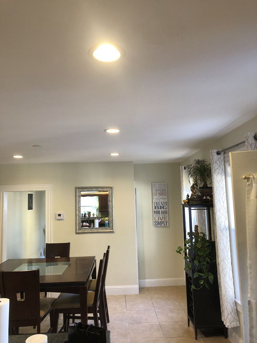 Dining room with table, chairs, mirror, and recessed lighting. Pale walls, tile floor, and a cabinet with plants.