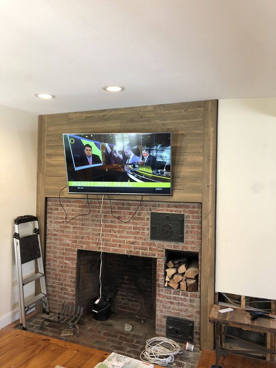 TV mounted above a brick fireplace, wires visible. Firewood in a nook. Brown, beige and white colors.