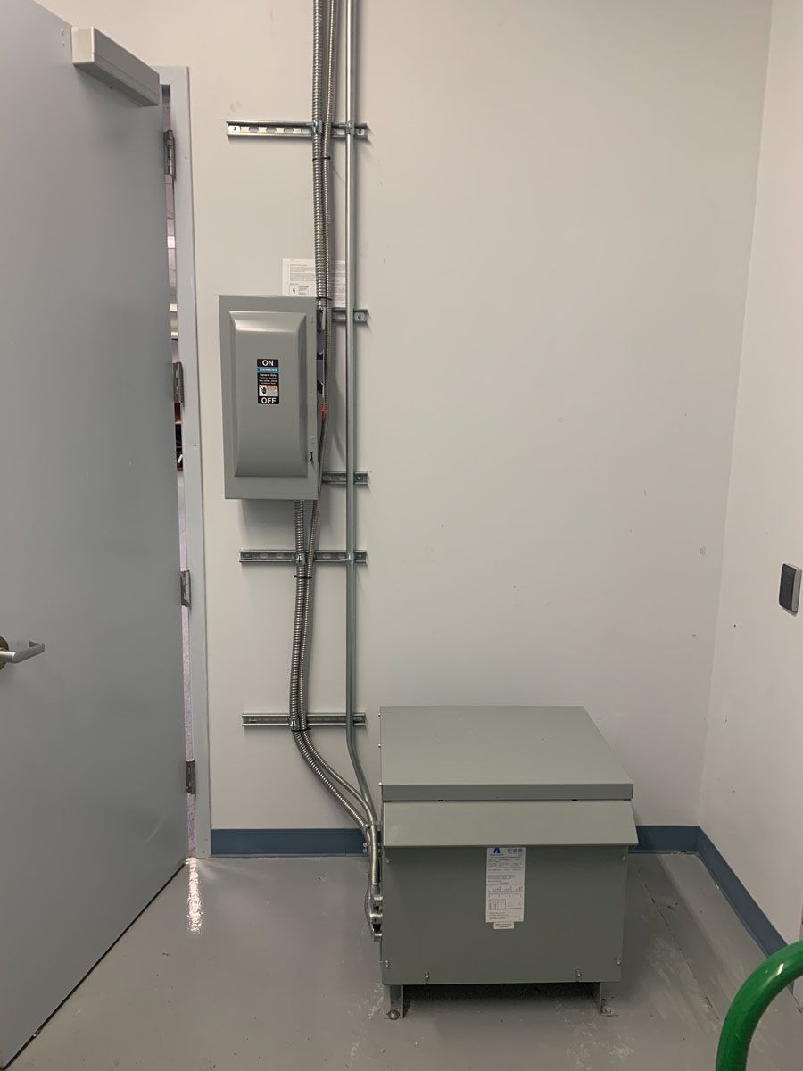 Electrical panel and transformer in a utility room. Gray door, white walls, and a gray floor.