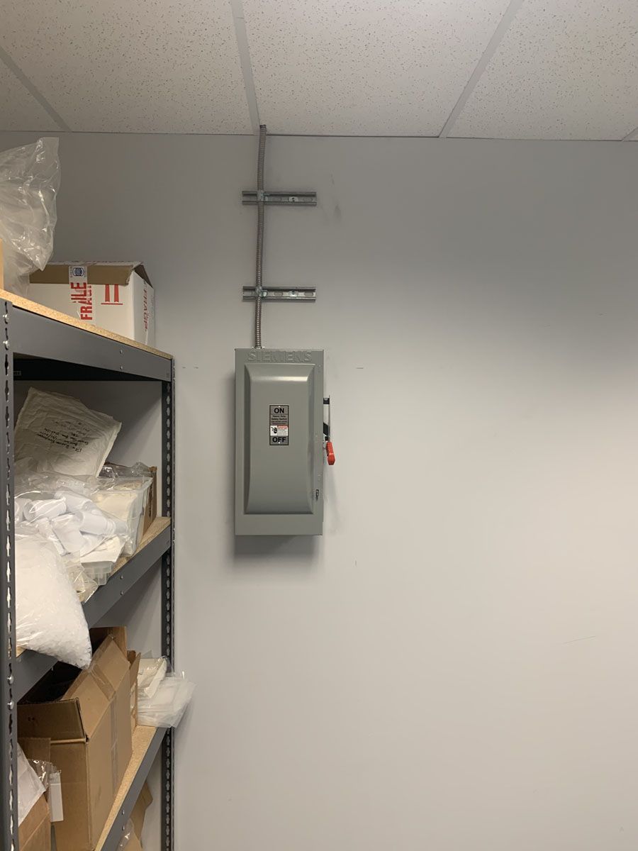 Gray electrical panel mounted on a white wall next to a metal shelf with boxes and supplies.