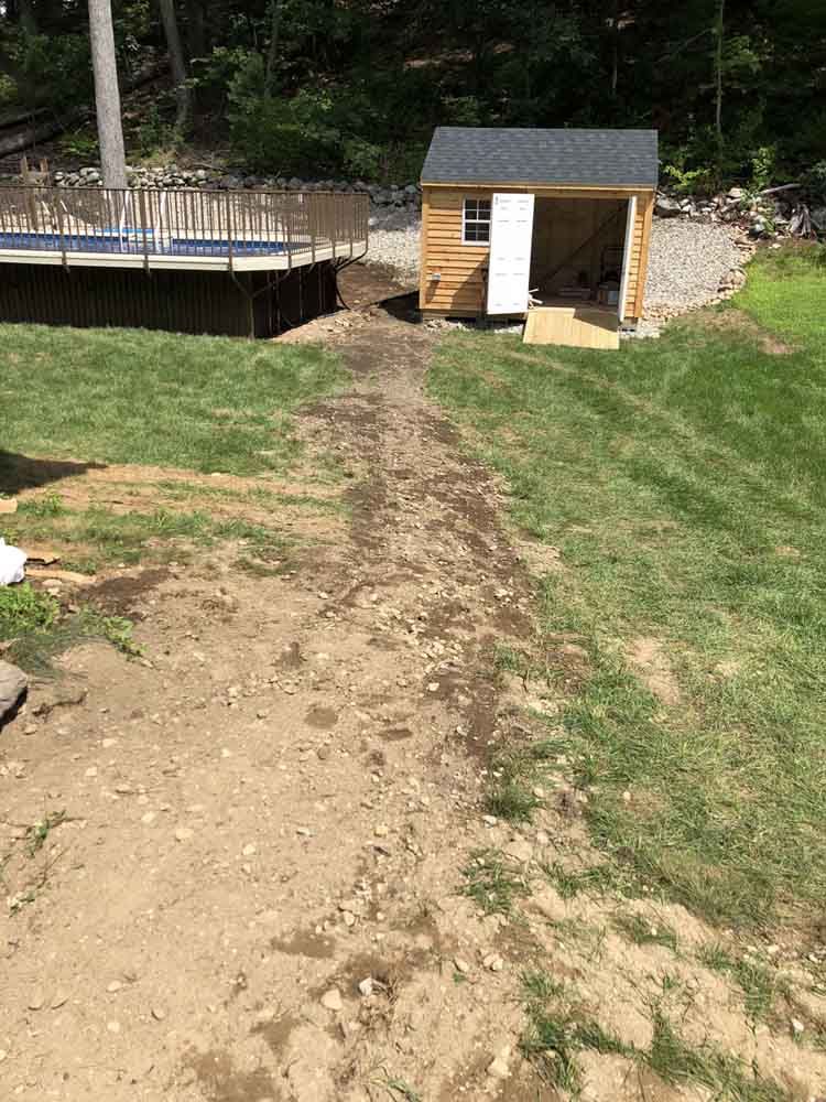 Dirt path leads from grassy yard to small shed near pool.