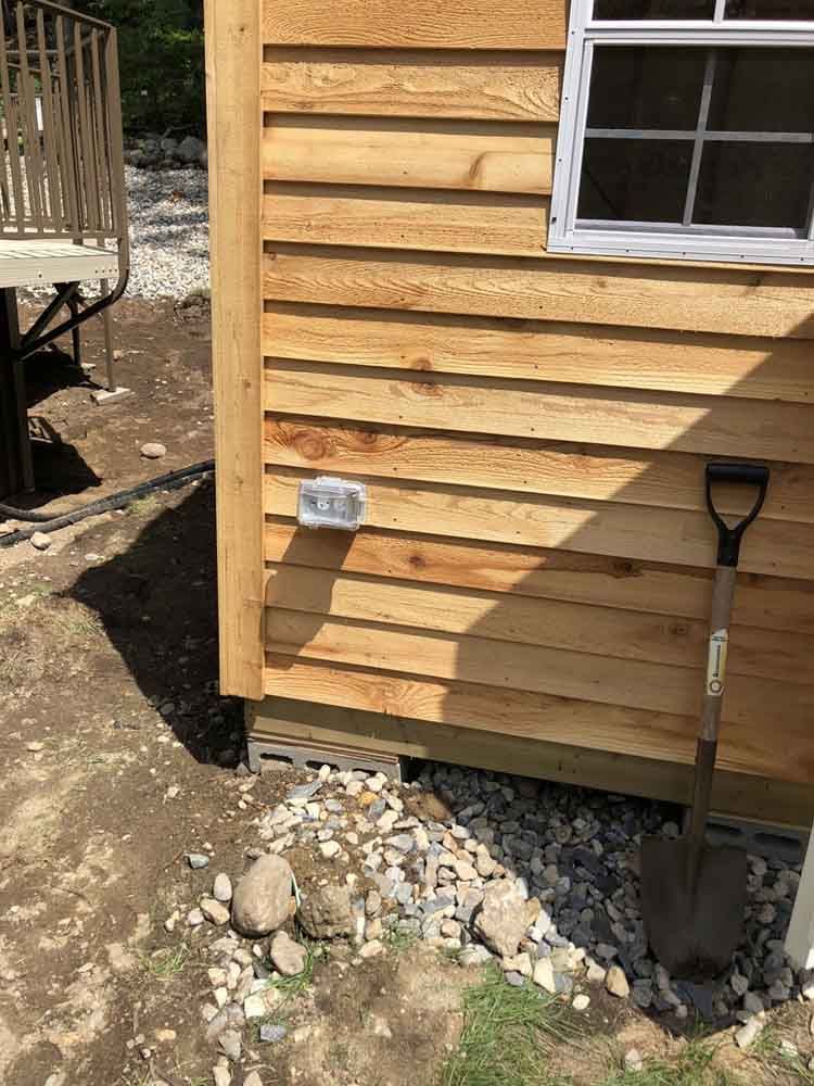 Exterior wall of a wooden structure with electrical outlet, window, and shovel; gravel and rocks on the ground.