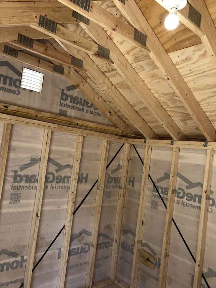 Interior of a wooden shed with a sloped ceiling, showing rafters, framing, and a small window.