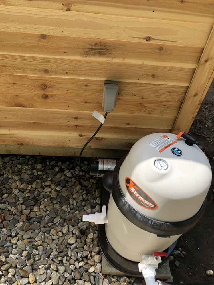 Spa equipment with electrical outlet against a wood siding wall, sitting on gravel.