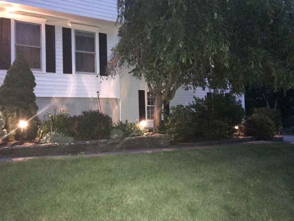 A white house with black shutters and landscaping illuminated by spotlights at dusk.