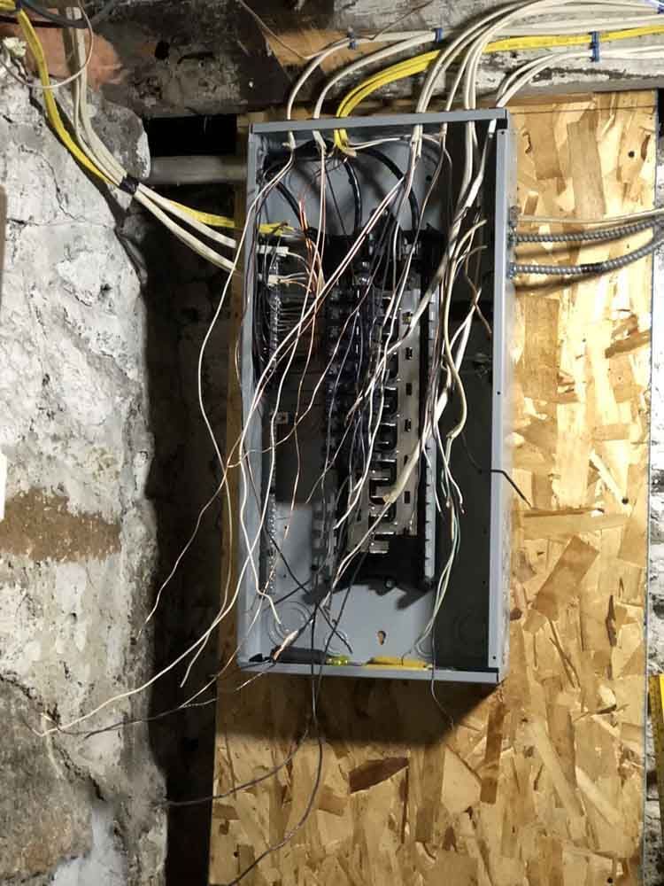 Electrical panel with numerous wires hanging from it, mounted on plywood and a concrete wall.