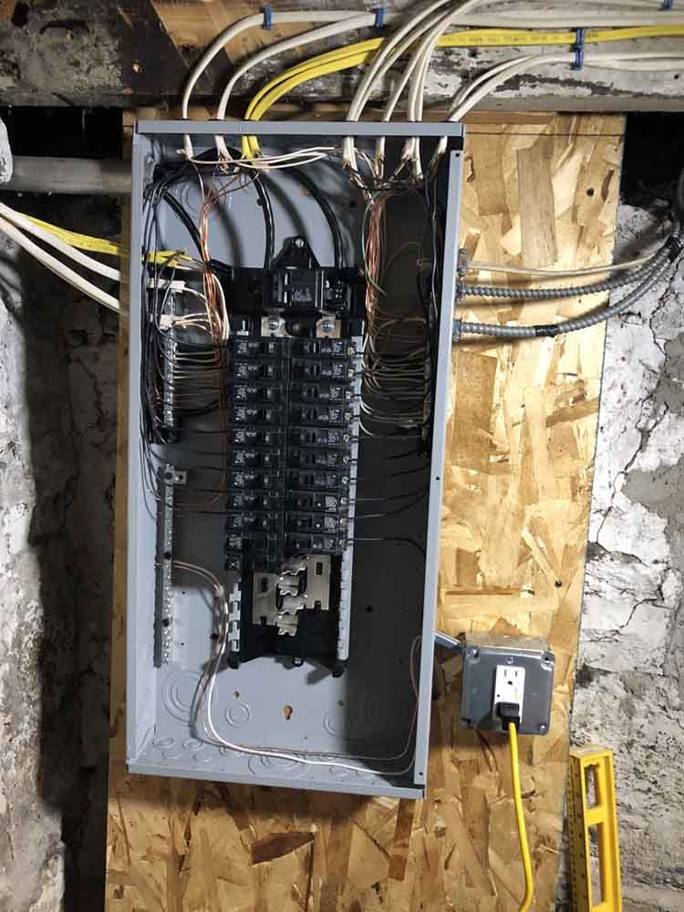 Electrical panel with wires, breakers, and a connected outlet on a wooden wall.