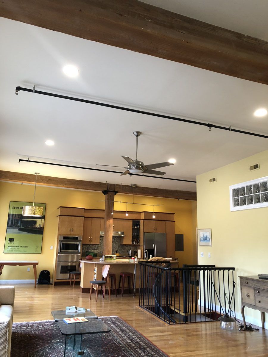 Open-concept loft with kitchen, dining, and living areas. Yellow walls, wooden beams, black pipe lighting, hardwood floors.