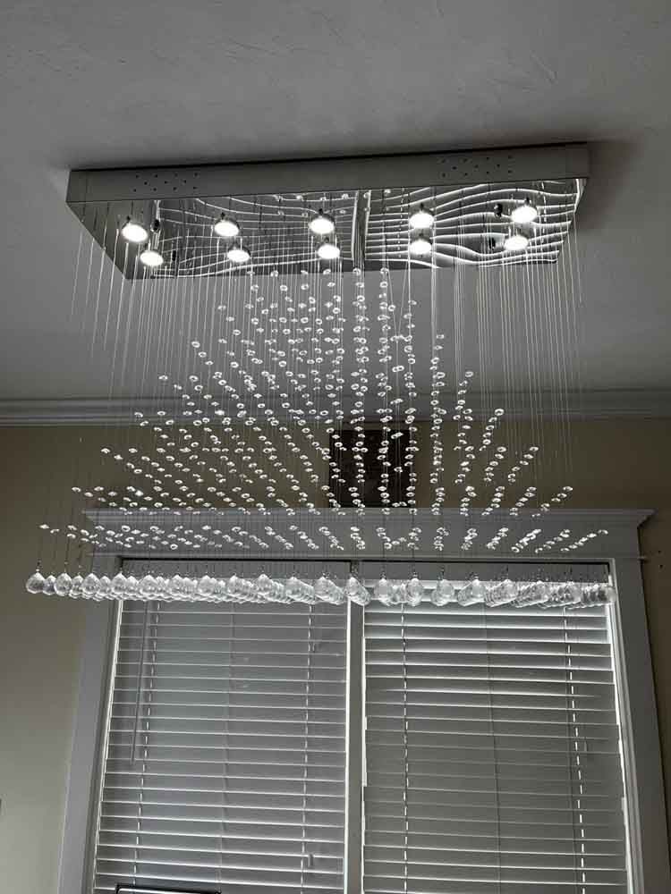 Modern ceiling light fixture with dangling crystal-like droplets, above a window with blinds.