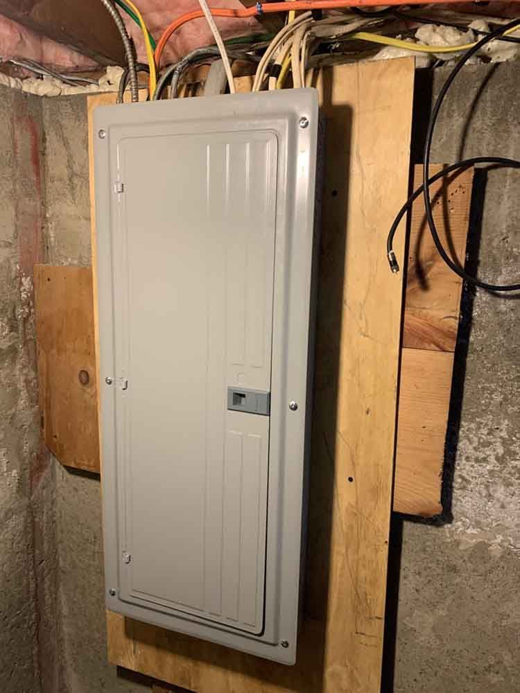 Electrical panel mounted on a wooden board in a basement, with wires running above.