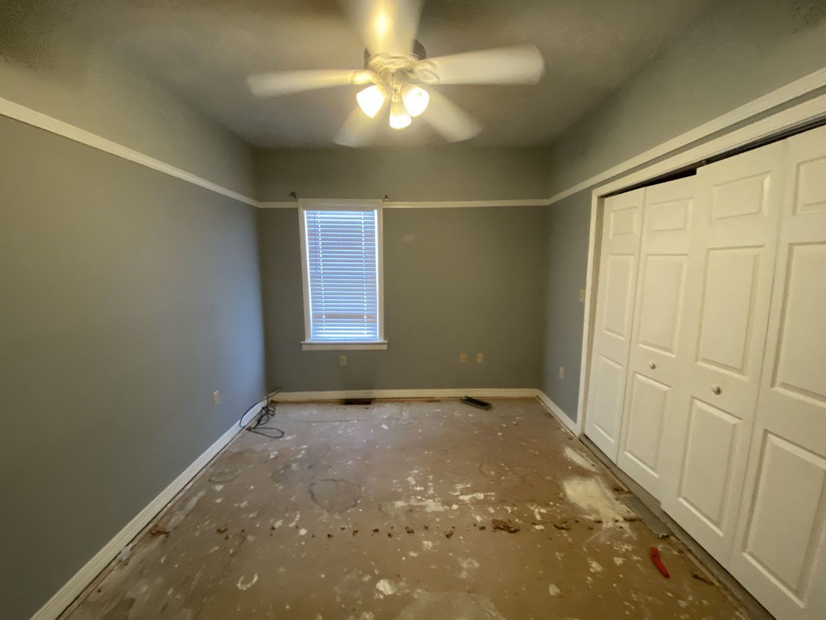 Empty gray room with white trim, closed closet doors, and a window. Damaged flooring visible.