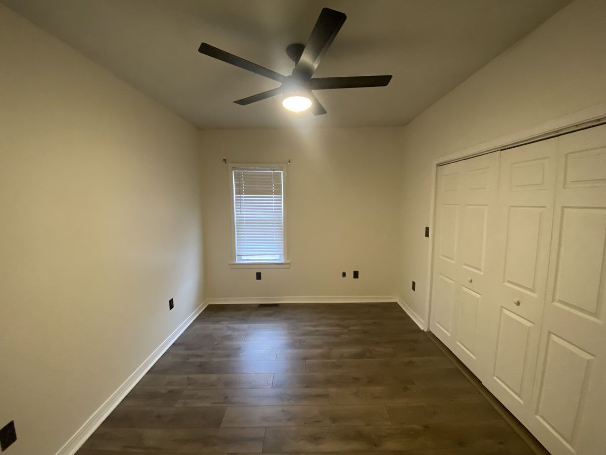 Empty bedroom with wood-look floor, white doors, window with blinds, ceiling fan, and tan walls.