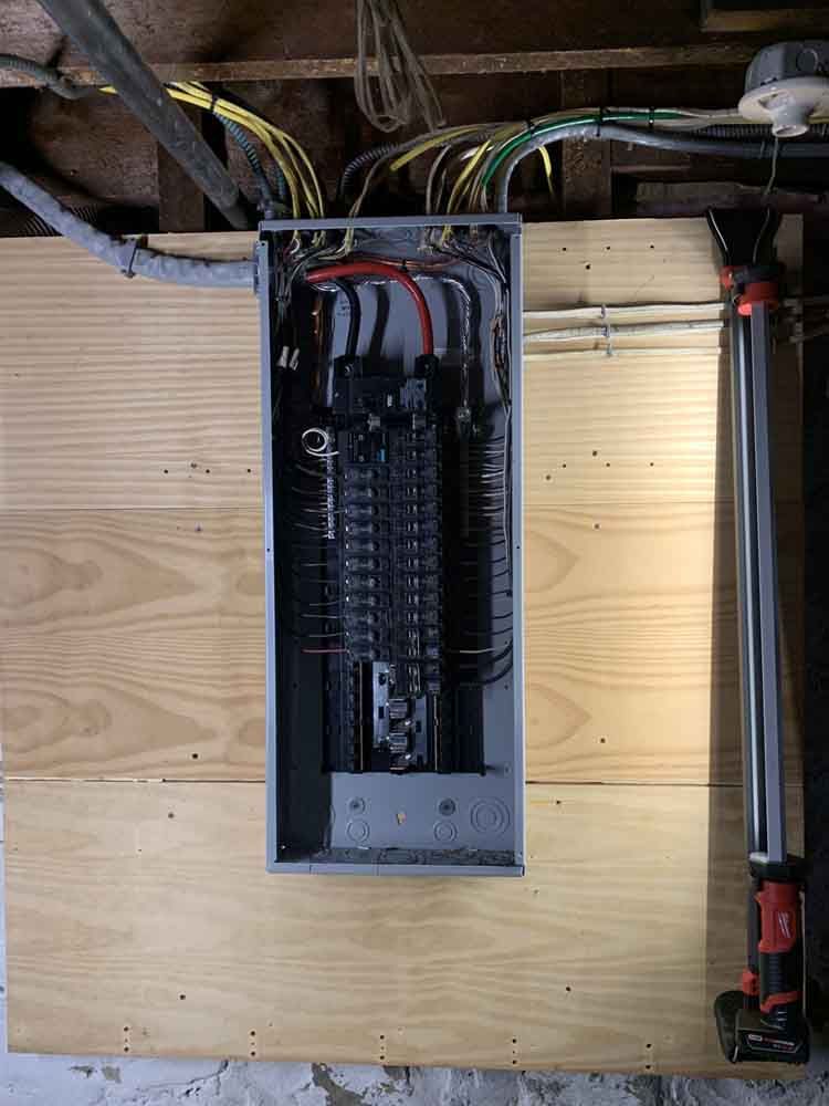 Electrical panel mounted on plywood in a basement, with wiring and a work light.