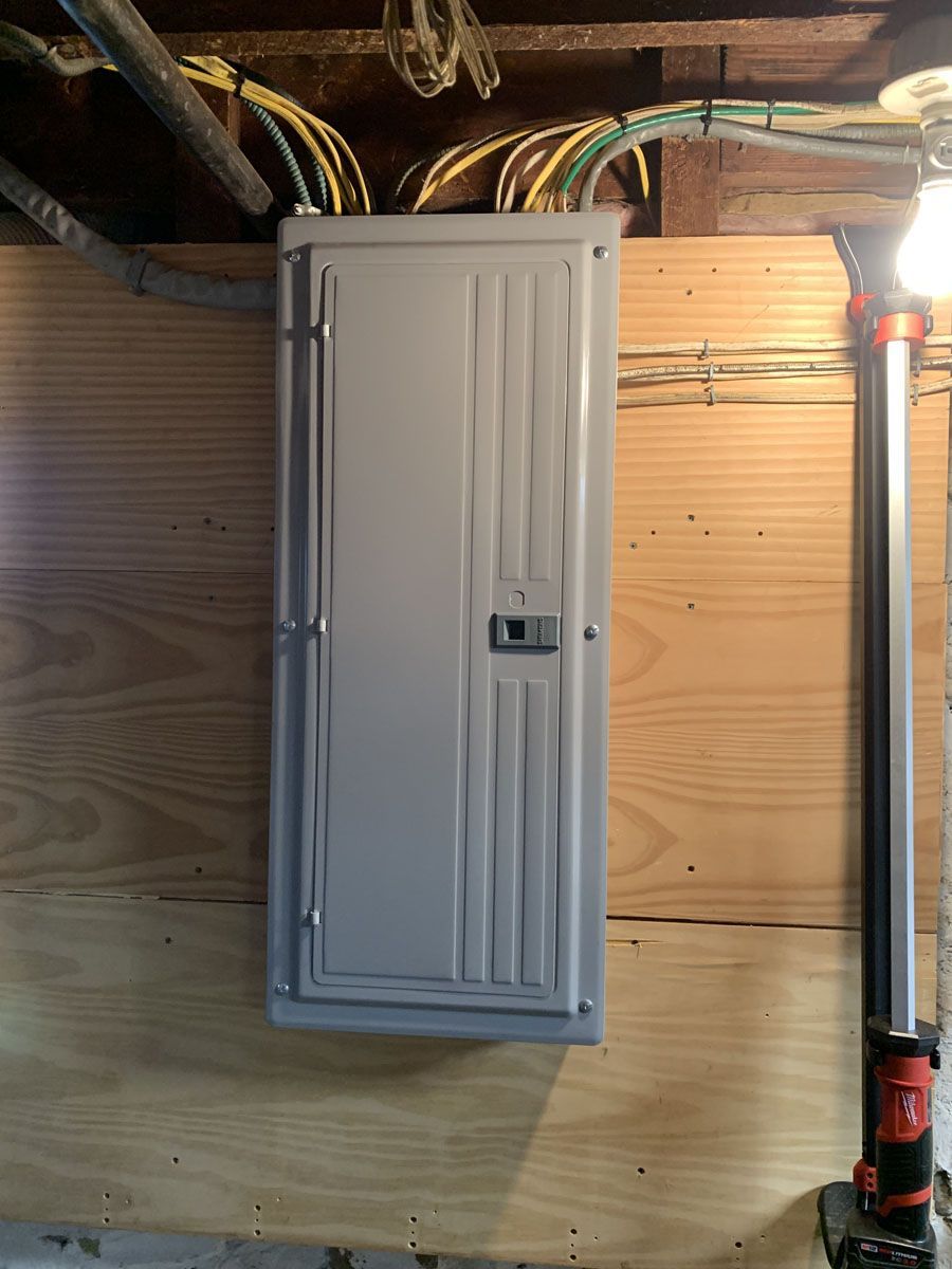 A gray electrical panel mounted on a plywood wall in a basement. Wiring and a work light are also present.