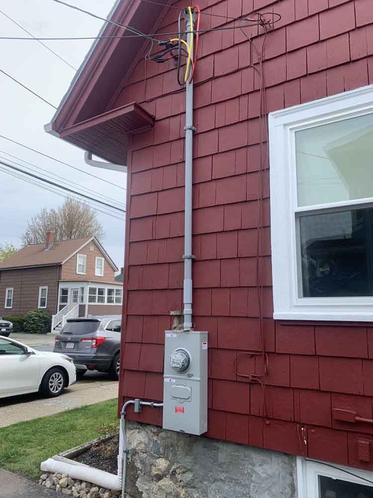 Gray electrical box and conduit on red house siding, with power lines overhead.