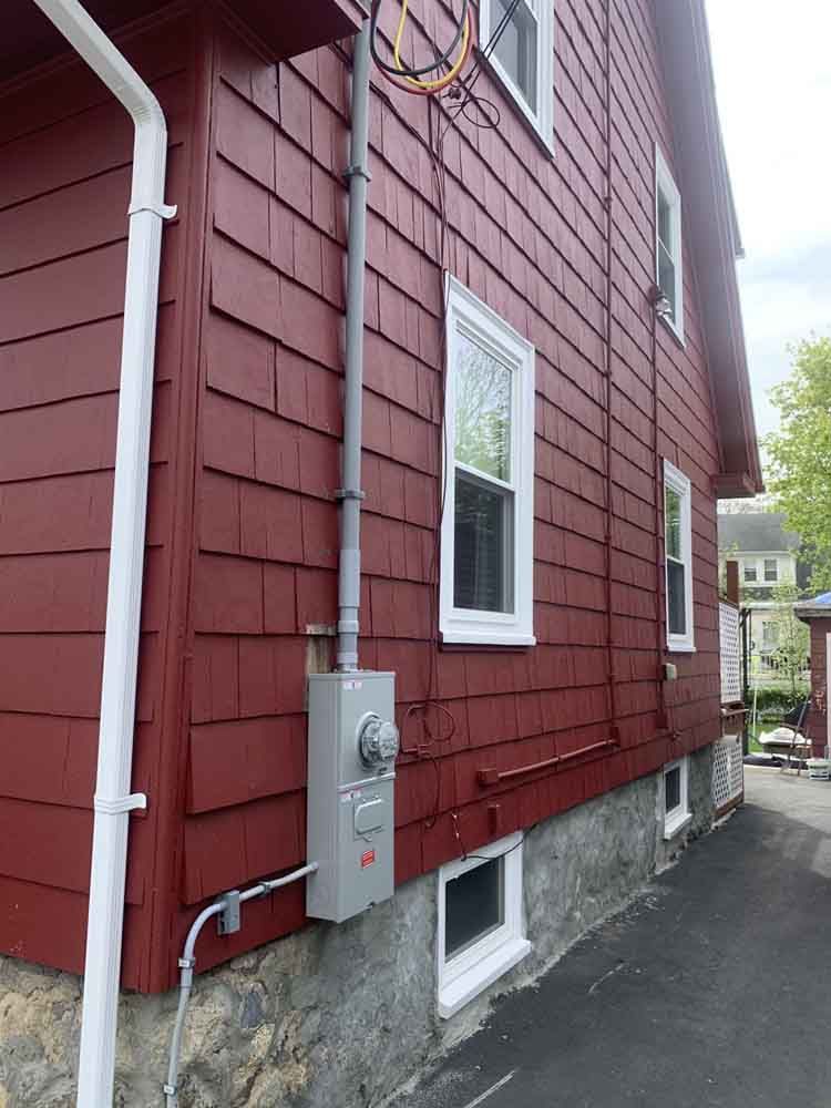 Red-sided house with a gray electrical meter and conduit. White trim, stone foundation, and asphalt driveway.