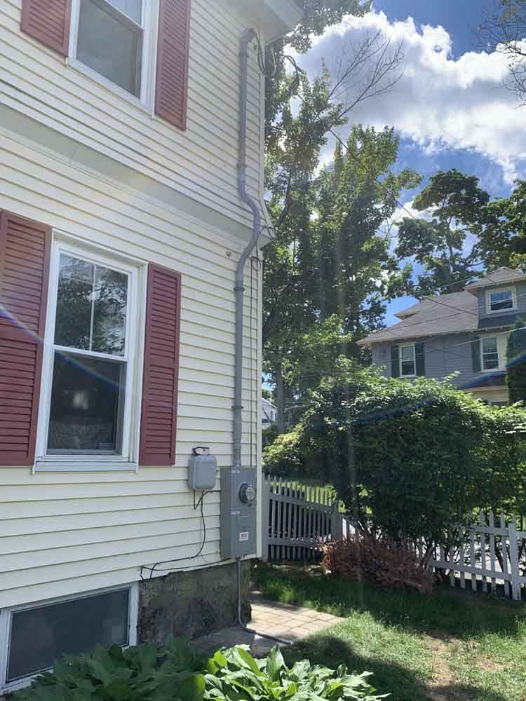 Beige house with red shutters, electrical box, and conduit leading up to power lines; sunny day.