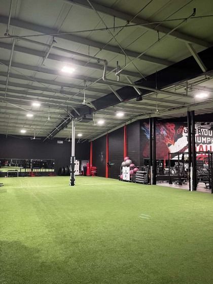 Spacious gym interior with green turf floor, black walls, and fitness equipment; a person in the background.