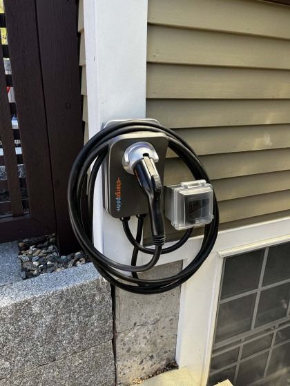 A black electric vehicle charger mounted on a house, cable coiled, next to an outdoor outlet.