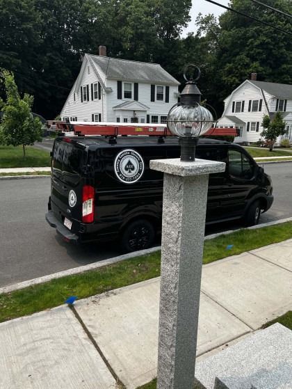 Black work van parked in front of a house, with a lamppost in the foreground, on a sidewalk.