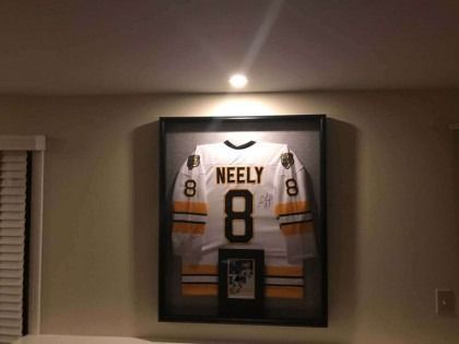 Framed hockey jersey, white with black and yellow stripes. Signed Neely #8 hangs on wall.
