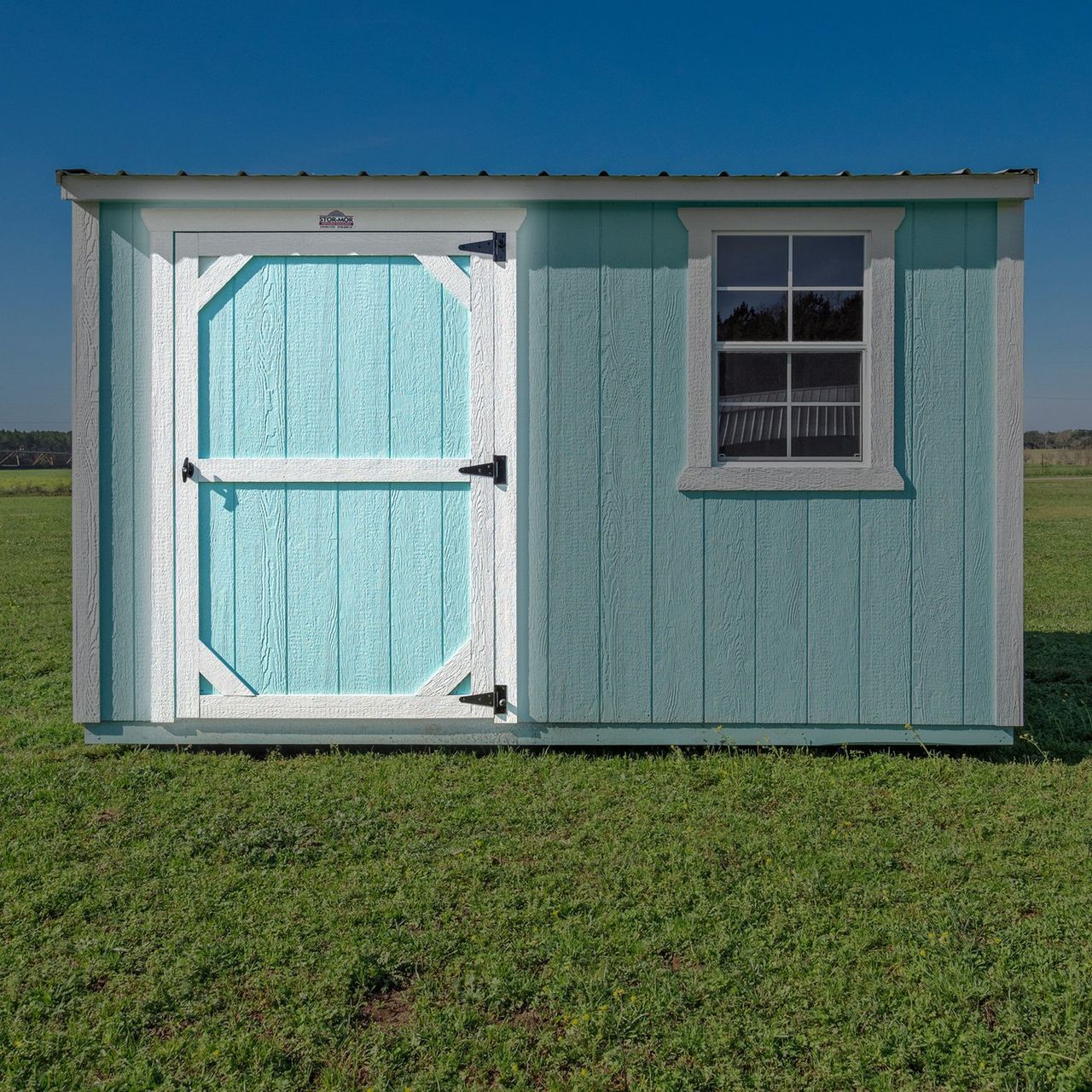 A blue and white shed with a window is sitting in the middle of a grassy field.