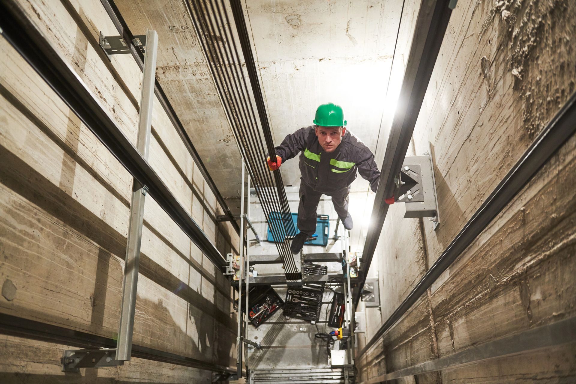 Un operaio con il casco verde scende lungo uno stretto pozzo industriale su una piattaforma sospesa.