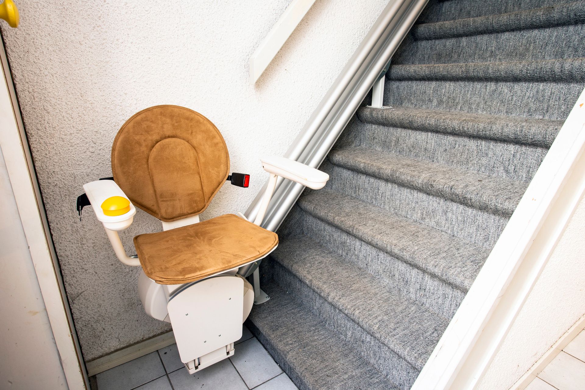 Montascale interno con sedile color beige accanto a una scala rivestita di moquette e corrimano bianco.