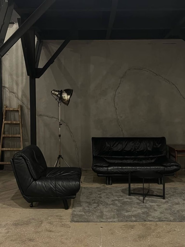 Black leather sofa and armchair in a dark, industrial-style room with a spotlight and small table.