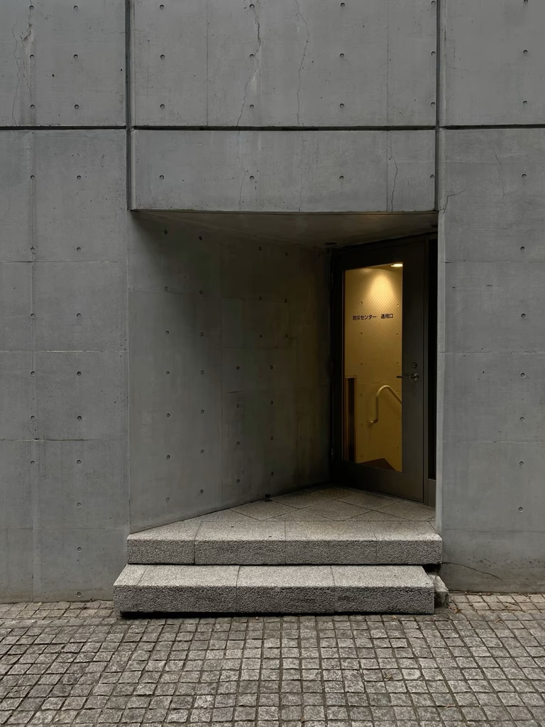 Concrete building entrance with steps leading to a glass door. Warm light glows from inside.