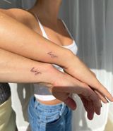 Two arms with small script tattoos crossed over a white top and blue jeans by a bright window